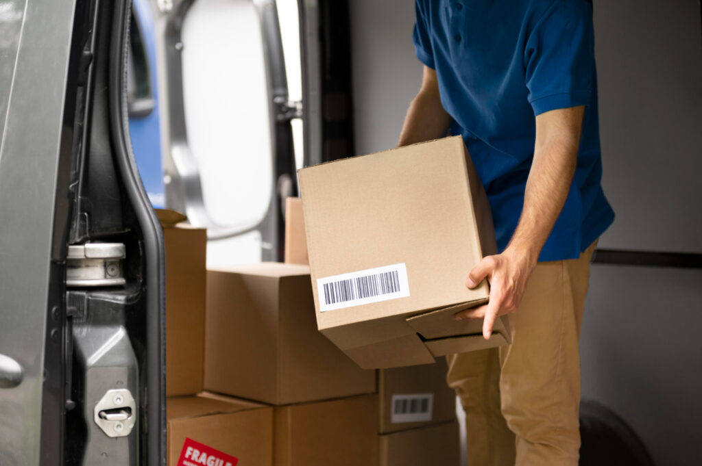 Amazon return pallets – A person carrying a cardboard box towards the open back of a delivery van filled with other boxes, suggesting the movement of Amazon returns.