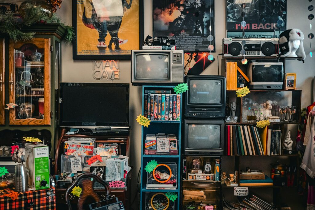 Shelves in a thrift store overflowing with vintage electronics like televisions and boomboxes, along with posters, books, and other miscellaneous goods that represent potential inventory for a reselling business.
